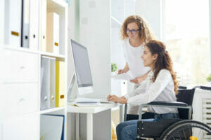 Two employees, one standing and one seated in a wheelchair, in a home care agency working on caregiver scheduling at the computer.