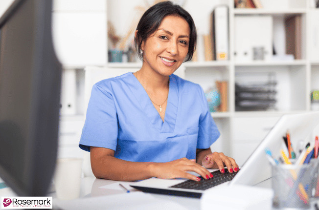 Woman in blue scrubs sitting at a desk working on a computer looking at the KPIs for her home care agency