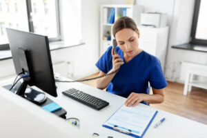 woman in blue scrubs sitting in front of a computer and talking on the phone while looking at patient paperwork