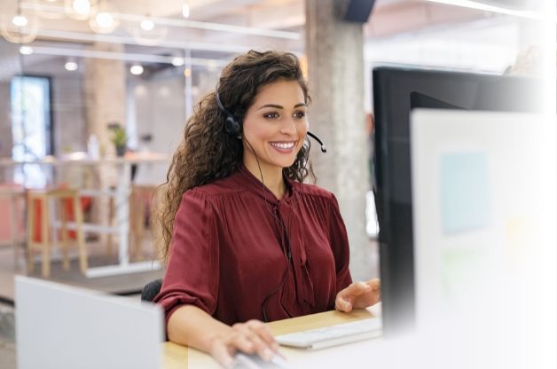 woman in rose-colored shirt with headphones on smiling at computer