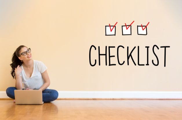 A woman sits on a wooden floor with a laptop, looking up thoughtfully at a wall that has “CHECKLIST” written on it with three checked boxes, symbolizing a completed Home Care Agency Year-End Checklist.