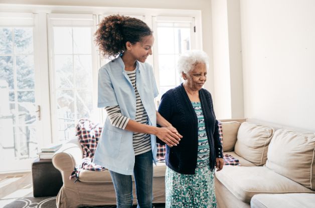 A smiling young female caregiver in a blue scrub top and striped shirt gently assisting an elderly woman to stand up from a sofa in a sunlit living room. The image conveys a sense of support, stability, and high-quality personal care.