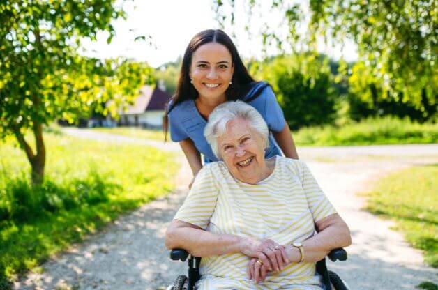 Female caregiver walking alongside a smiling female client in a wheelchair outdoors, enjoying time together in a bright, natural setting.