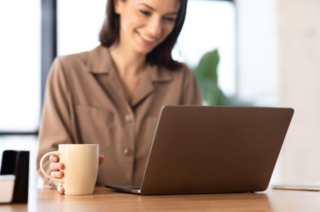 A smiling woman working on a laptop with a coffee mug, representing a home care agency owner researching Home Care GEO vs SEO strategies for 2026.