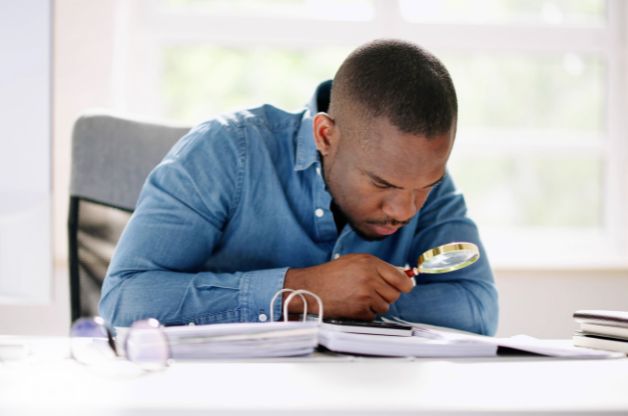 A focused professional using a magnifying glass to closely review paperwork and files, illustrating the detailed nature of home care audit preparedness and internal compliance reviews.