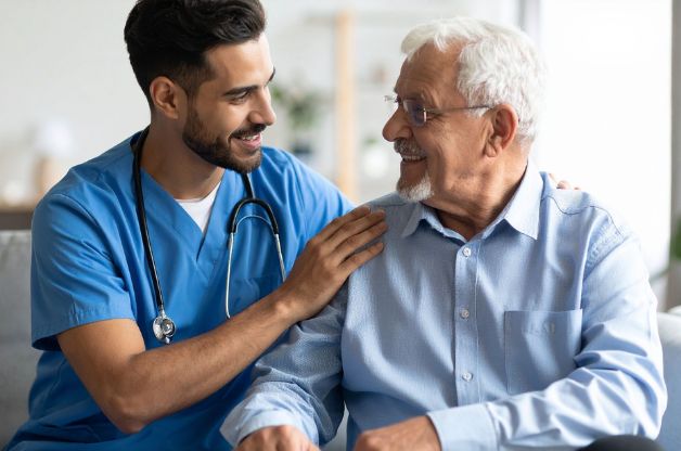 A smiling home caregiver in blue scrubs sits beside an older adult client, gently placing a hand on his shoulder in a moment of connection and trust.